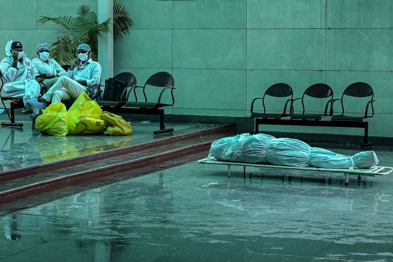 Profissionais de saúde descansam próximo a corpo de vítima da Covid-19 em crematório de Nova Déli, na Índia
