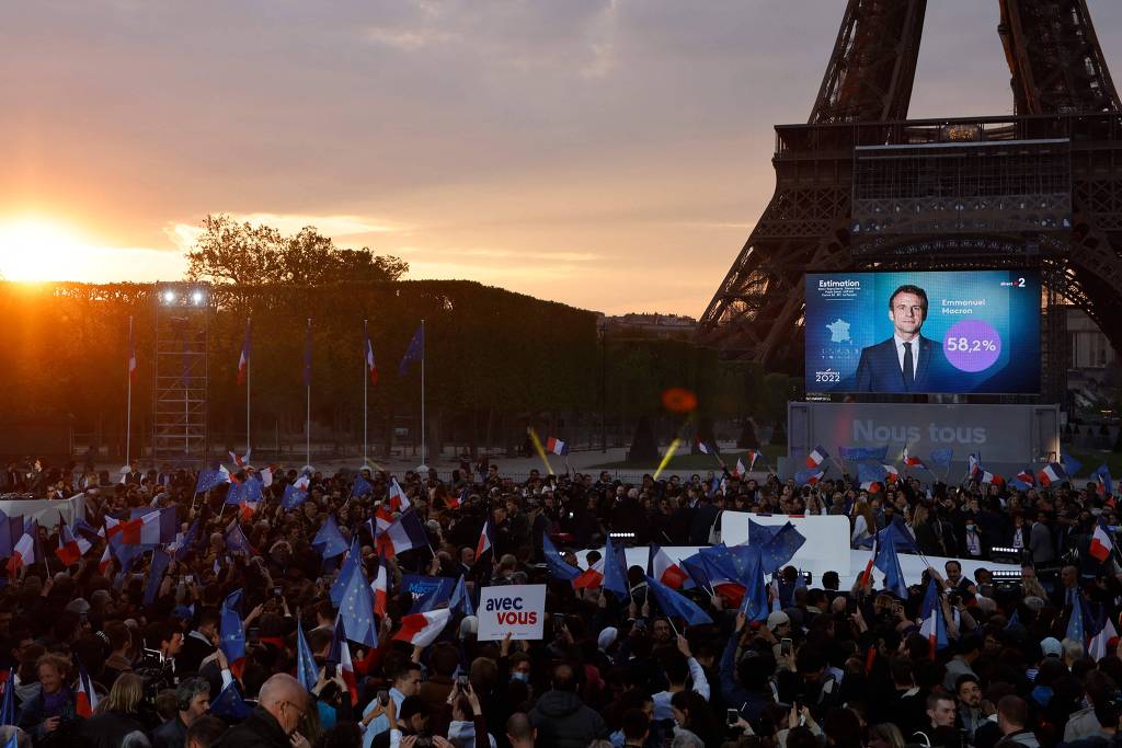 Apoiadores de Emmanuel Macron celebram divulgação das projeções, em telão, indicando a reeleição do presidente, no campo de Marte, em frente à Torre Eiffel, em Paris, neste domingo